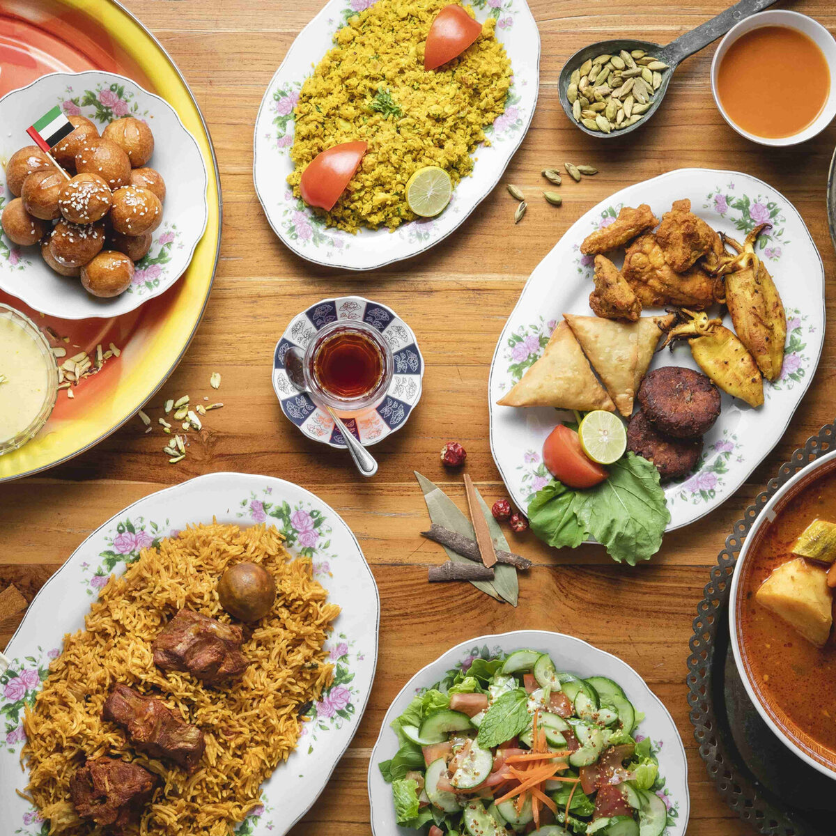 Emirati dishes on a wooden table seen from above: rice, stews, salad, samosas and luqaimat